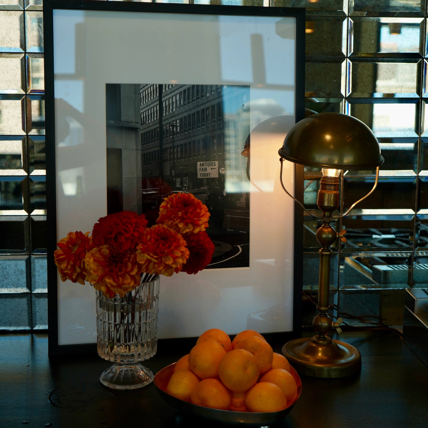 Decorative setup with flowers, oranges, and a framed picture on a reflective surface.