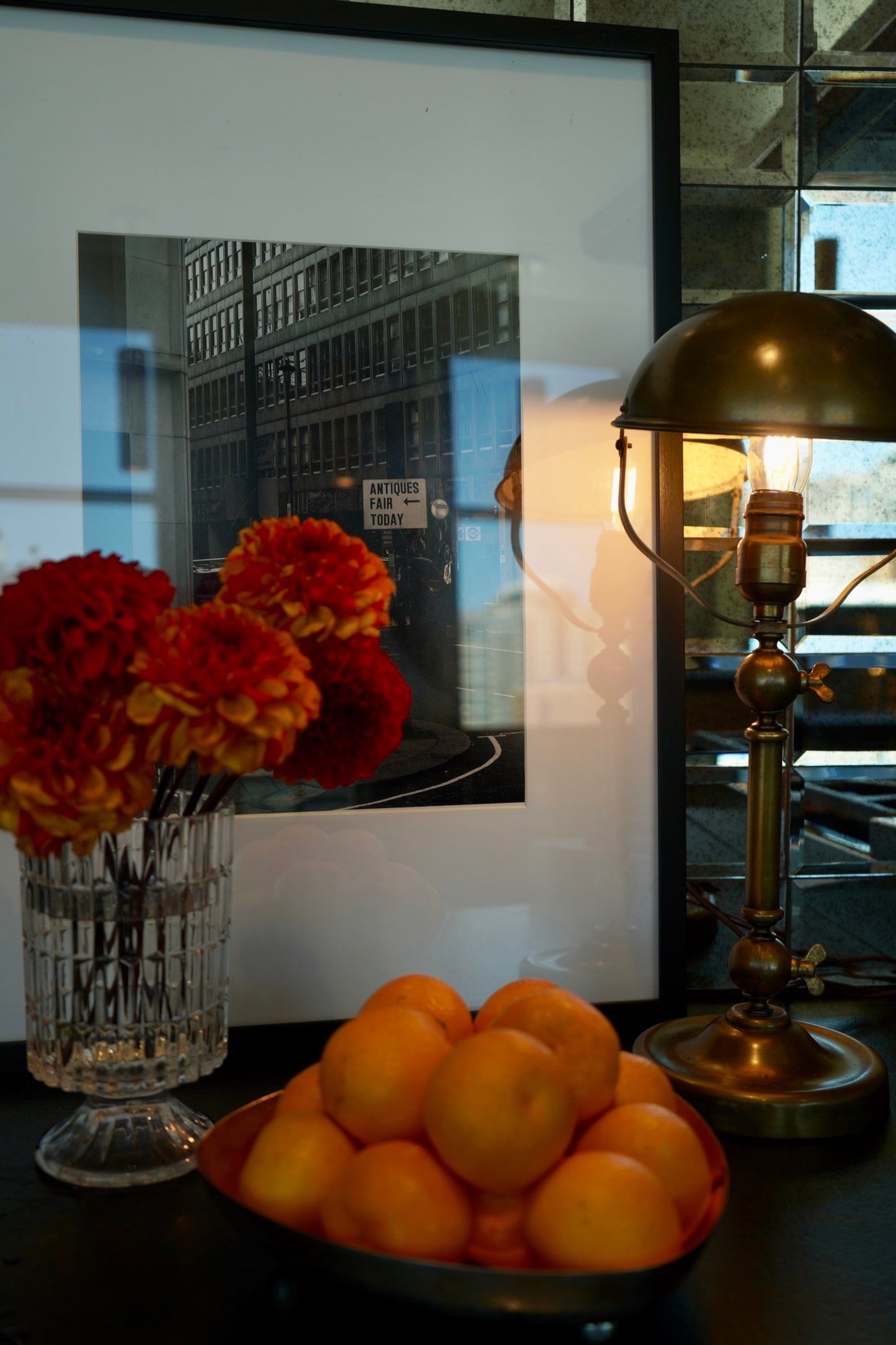 Bowl of oranges with a vase of flowers and a lamp on a reflective surface
