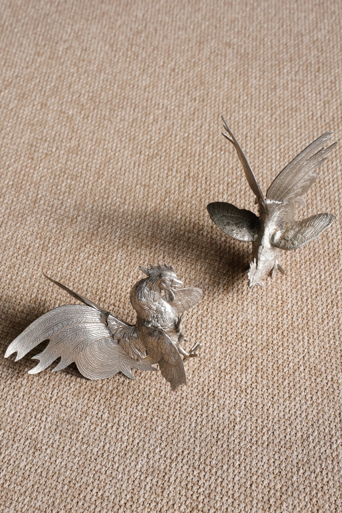 Two small metallic bird figurines with spread wings, placed on a textured beige fabric background.