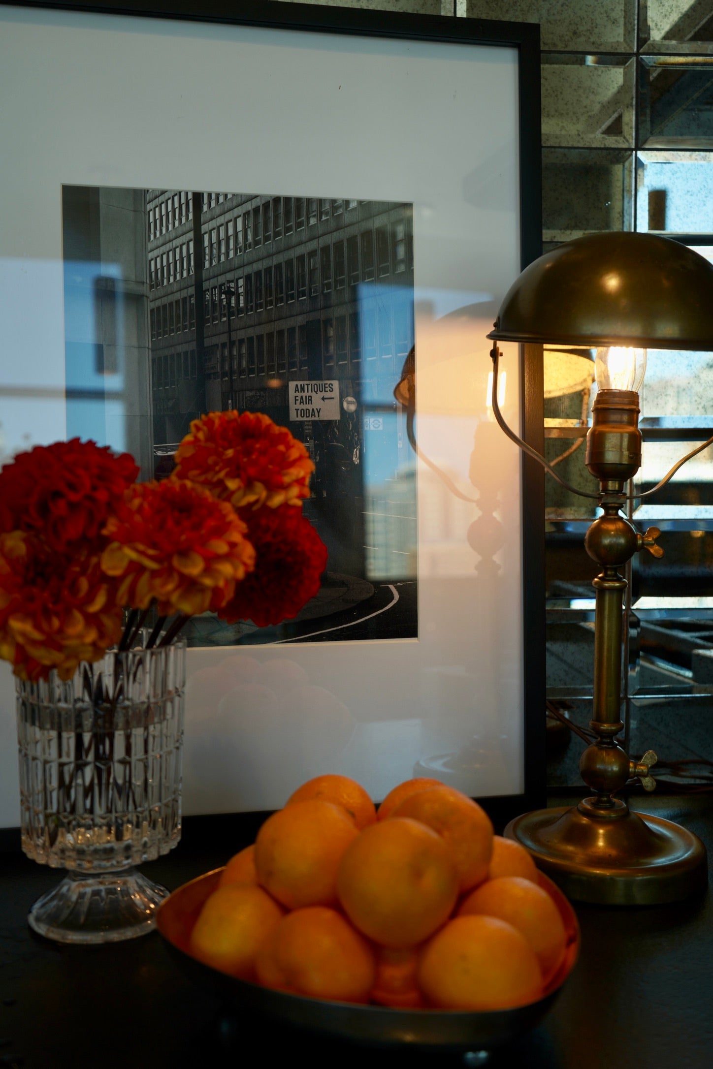 Bowl of oranges with a vase of flowers and a lamp on a reflective surface