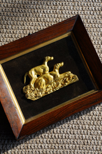 A square framed decorative piece featuring a raised gold figure mounted on a black background, set within a dark wood frame with a gold inner border, photographed on a textured beige surface.